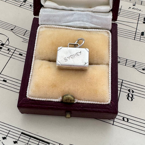 Silver vintage suitcase charm with 'SYDNEY' engraved on it in a jewelry box with music sheet background