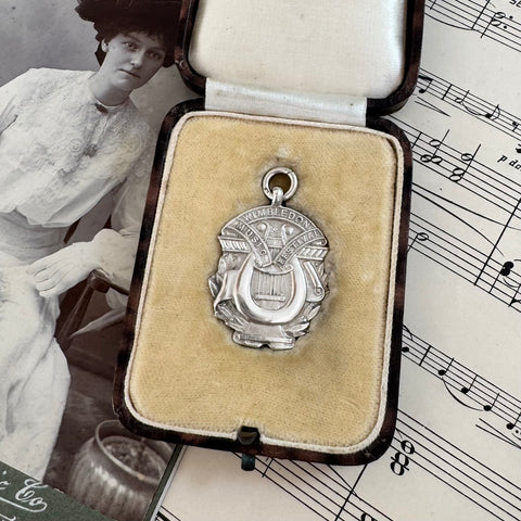 Vintage wimbledon Music Festival silver medallion in an open jewelry box on sheet music with a vintage-style woman in the background.