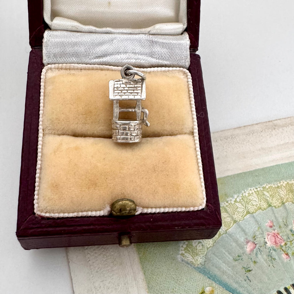 Vintage silver wishing well charm in an open jewelry box on a textured surface