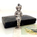 Vintage sterling silver needle case in the form of a boy holding a bunch of flowers. There is a vintage postcard and jewellery box in the background.
