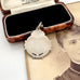 A vintage sterling silver watch fob pendant with a scalloped border and small engraved text, displayed in front of an open vintage wooden jewelry box on top of an old sepia portrait photograph.
