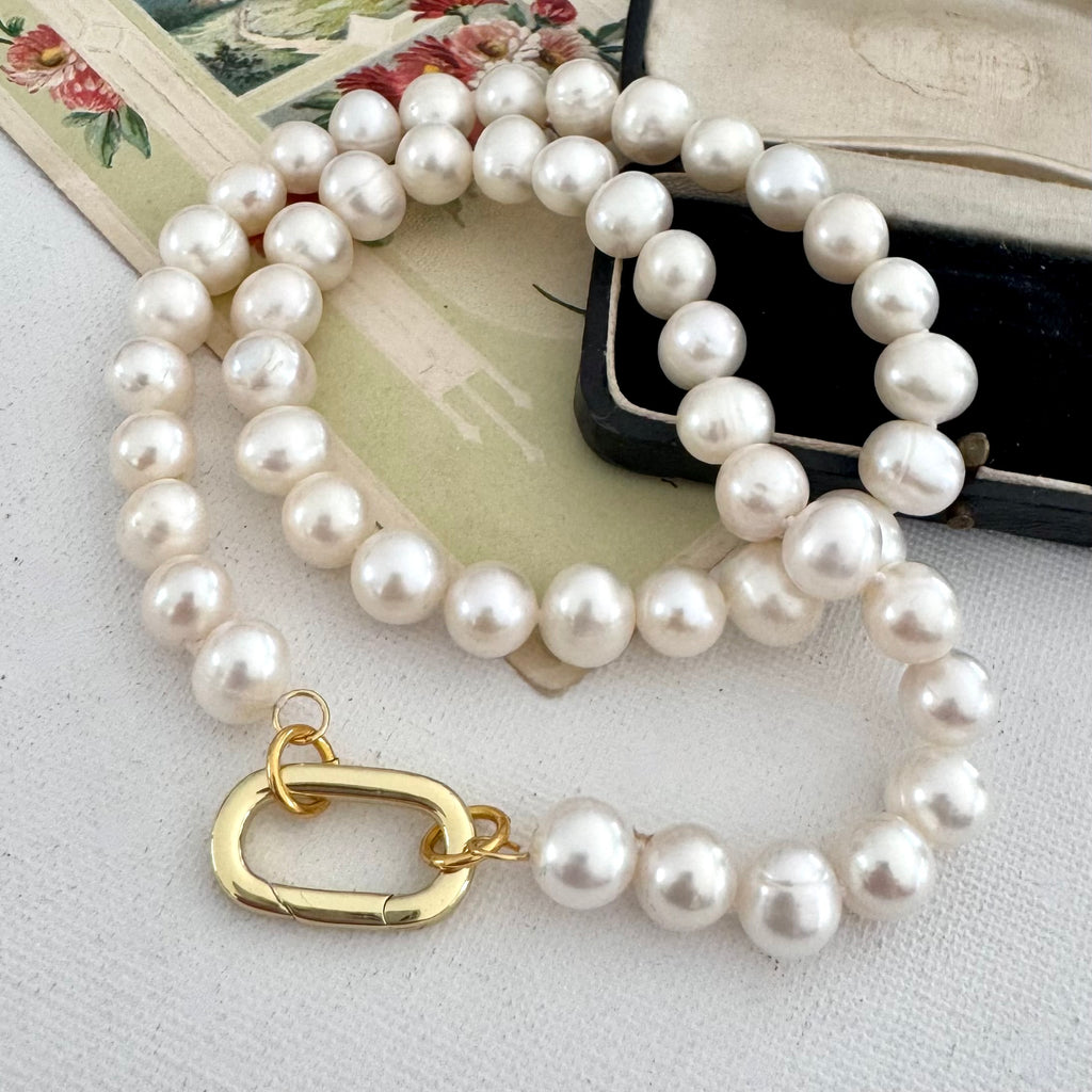 A strand of lustrous white freshwater pearls arranged in a loop with a gold clasp, photographed on a light fabric surface with vintage paper and a black case as styled props.