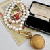 A vintage-style pearl strand necklace with a gold clasp displayed next to an open black velvet jewelry box and a gold pocket watch, arranged on an antique floral card over a white fabric background.
