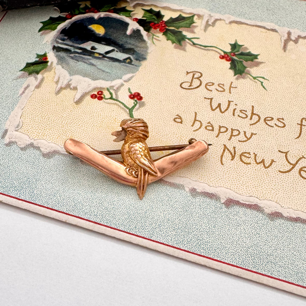 Decorative card with a gold brooch featuring a kookaburra, holly leaves, and berries on a textured surface.