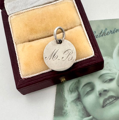 Silver pendant with 'M. B.' engraving in a jewelry box with a vintage photo in the background.