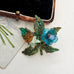 A vintage-style brooch featuring a green glitter-leaf design with a blue flower, a light green stone, and a brown stone, displayed on a light surface next to a black velvet case.