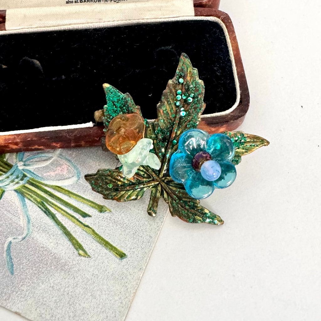 A vintage-style floral  brooch featuring green glitter leaves, a blue flower with a light blue center, and a small amber-colored flower, displayed next to an open black jewelry box on a white surface.