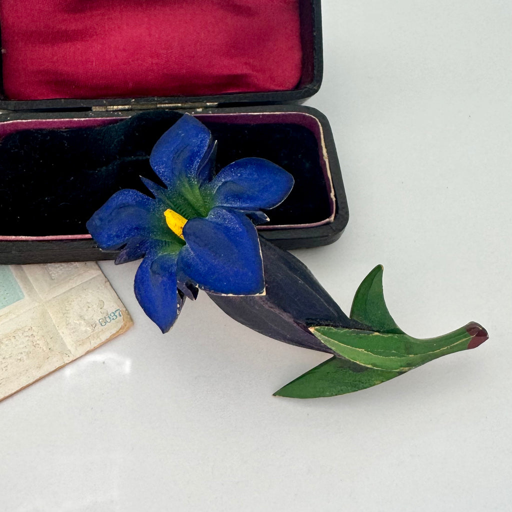 Vintage Celluloid Gentian blue flower brooch with green leaves on a white surface next to an open jewelry box.