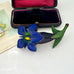 Vintage Celluloid Gentian blue flower brooch with green leaves on a white surface next to an open jewelry box.