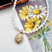 A white pearl beaded necklace with an oval rolled gold engraved vintage locket, displayed diagonally over a vintage floral card featuring yellow daisies.