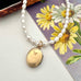 A pearl strand necklace with a gold oval locket pendant, displayed on a floral card over a light surface. The locket is engraved with 'Rolled GOLD'.