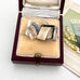 A miniature silver book-shaped pendant with a silver ring clasp, displayed inside an open vintage burgundy velvet jewelry box with a cream lining. The box is placed on a white surface next to a decorative card featuring lily of the valley flowers.