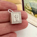 A small silver book-shaped pendant with a cross on the front, held between fingers to show its size. The pendant has engraved decorative borders and a silver bail at the top.