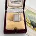 A vintage silver bible charm presented in an open dark red velvet jewelry box with cream lining. The watch has a rectangular body with decorative geometric borders on the front and a small top clasp.