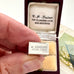 A hand holds a small silver emergency money charm engraved with 'IN EMERGENCY BREAK GLASS' in front of an open vintage dark red presentation box with cream lining and printed address text.