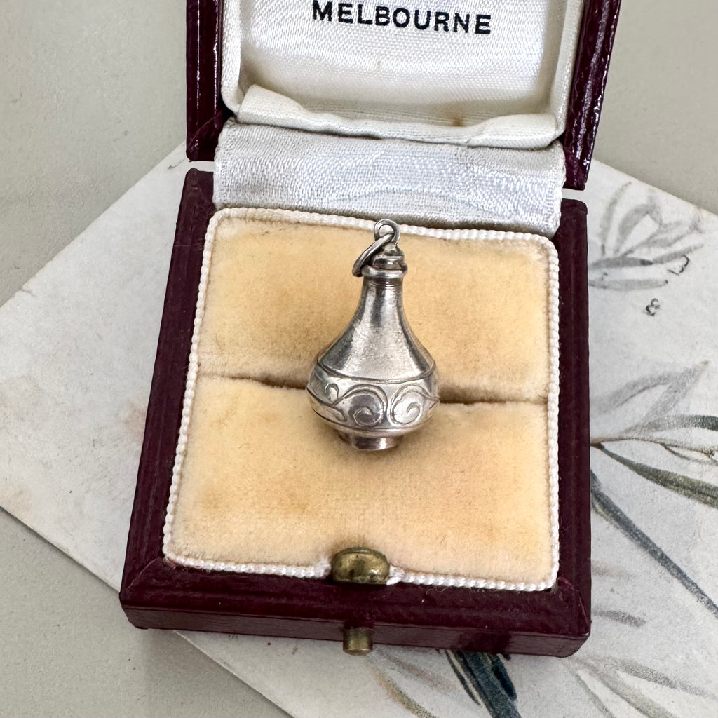 A vintage vase or urn charm with engraved swirl patterns sits in an open vintage jewelry box lined with cream velvet. The box has a dark burgundy exterior and the inside lid is lined with white fabric printed with the word 'MELBOURNE'.