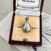 A vintage vase or urn charm with engraved swirl patterns sits in an open vintage jewelry box lined with cream velvet. The box has a dark burgundy exterior and the inside lid is lined with white fabric printed with the word 'MELBOURNE'.