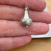 A small silver pendant shaped like a decorative bottle or teardrop with engraved swirl patterns, held between a person's fingers over a light wood surface.