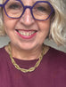 A close-up of a smiling woman with gray hair wearing purple geometric eyeglasses and a chunky gold chain necklace, paired with a burgundy top.