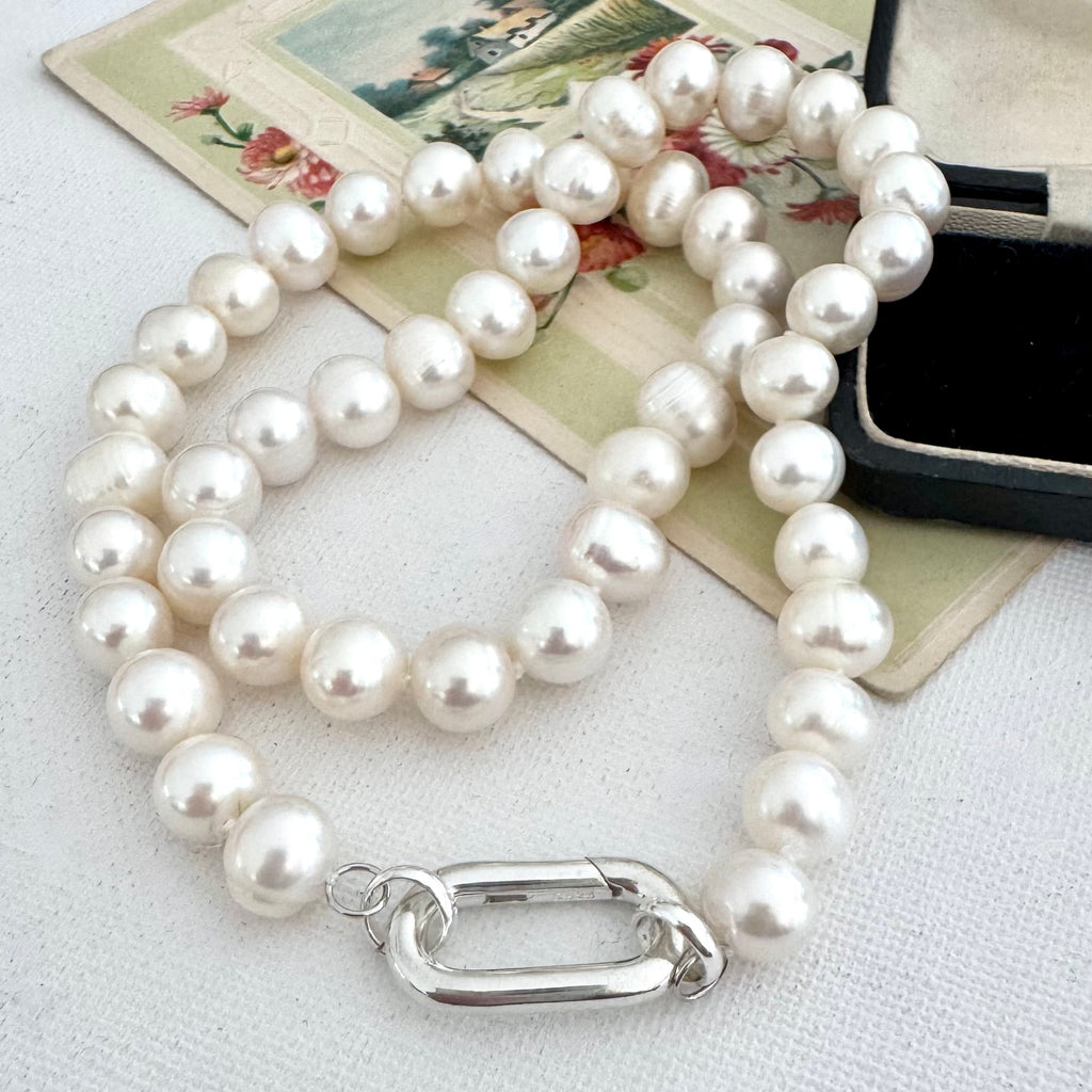 A  pearl necklace with a silver clasp, displayed on a textured white fabric alongside a vintage floral card and a black jewelry box.