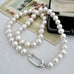 A strand of lustrous white pearls arranged in loops with a polished silver-toned oval clasp, photographed on a light fabric surface with a vintage floral card and a black jewelry box as props.